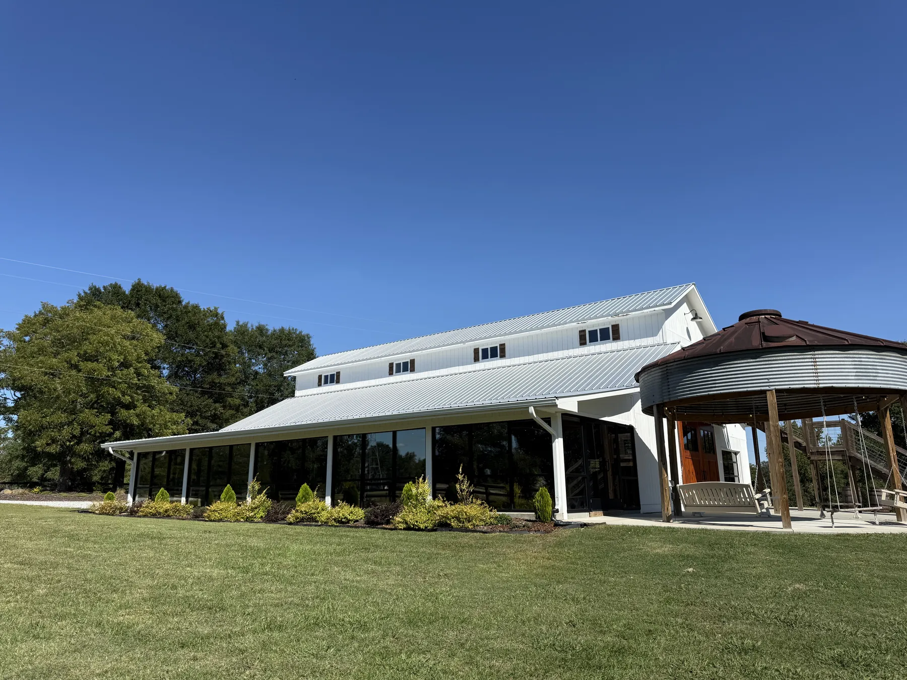 The white barn with its glass-enclosed reception space along the side, landscaped beds, and the silo swings beside it under a clear blue sky