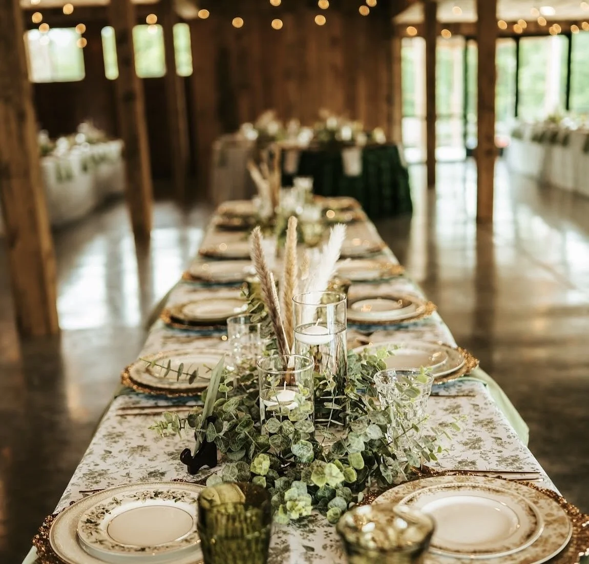 A long farm table with eucalyptus garland, pampas grass, floating candles and gold-rimmed china beneath the barn string lights