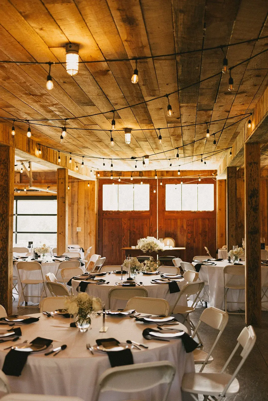 Round tables with white linens and floral centerpieces beneath string lights and wood beams in the barn