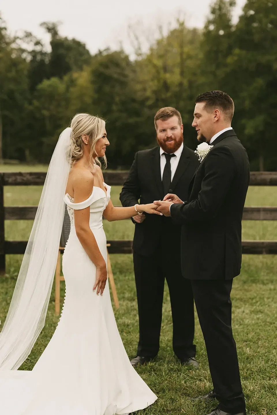 A bride and groom holding hands during their ceremony on the lawn, white florals and pasture fence behind