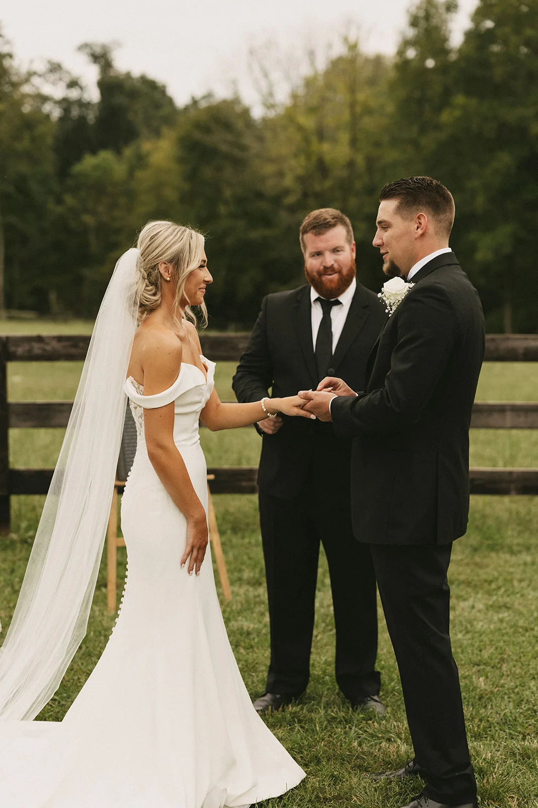 A bride and groom holding hands during their ceremony on the lawn, white florals and pasture fence behind