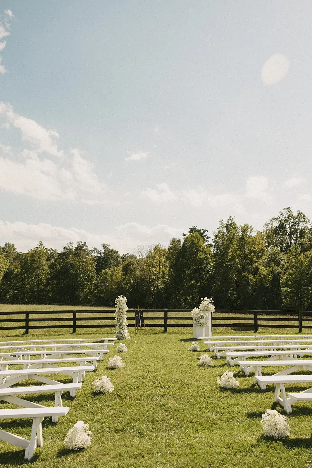 Rows of white benches on the ceremony lawn under a blue sky, with white floral pedestals and trees beyond