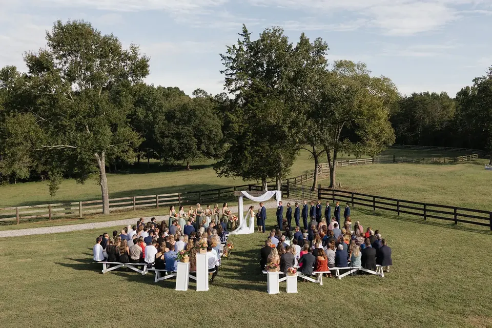 Wedding guests seated on white benches on the ceremony lawn, the couple beneath a draped arch, with pasture fence and trees beyond
