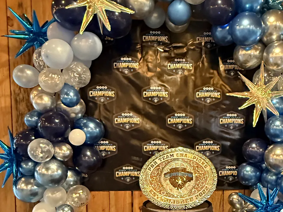 A balloon arch and championship backdrop framing a trophy buckle on a whiskey barrel