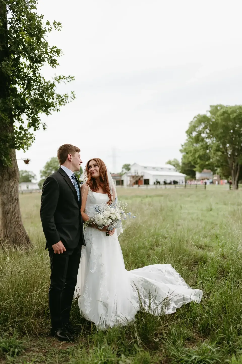 A bride and groom standing in tall grass beneath an oak tree, with the white barn and wedding guests across the pasture