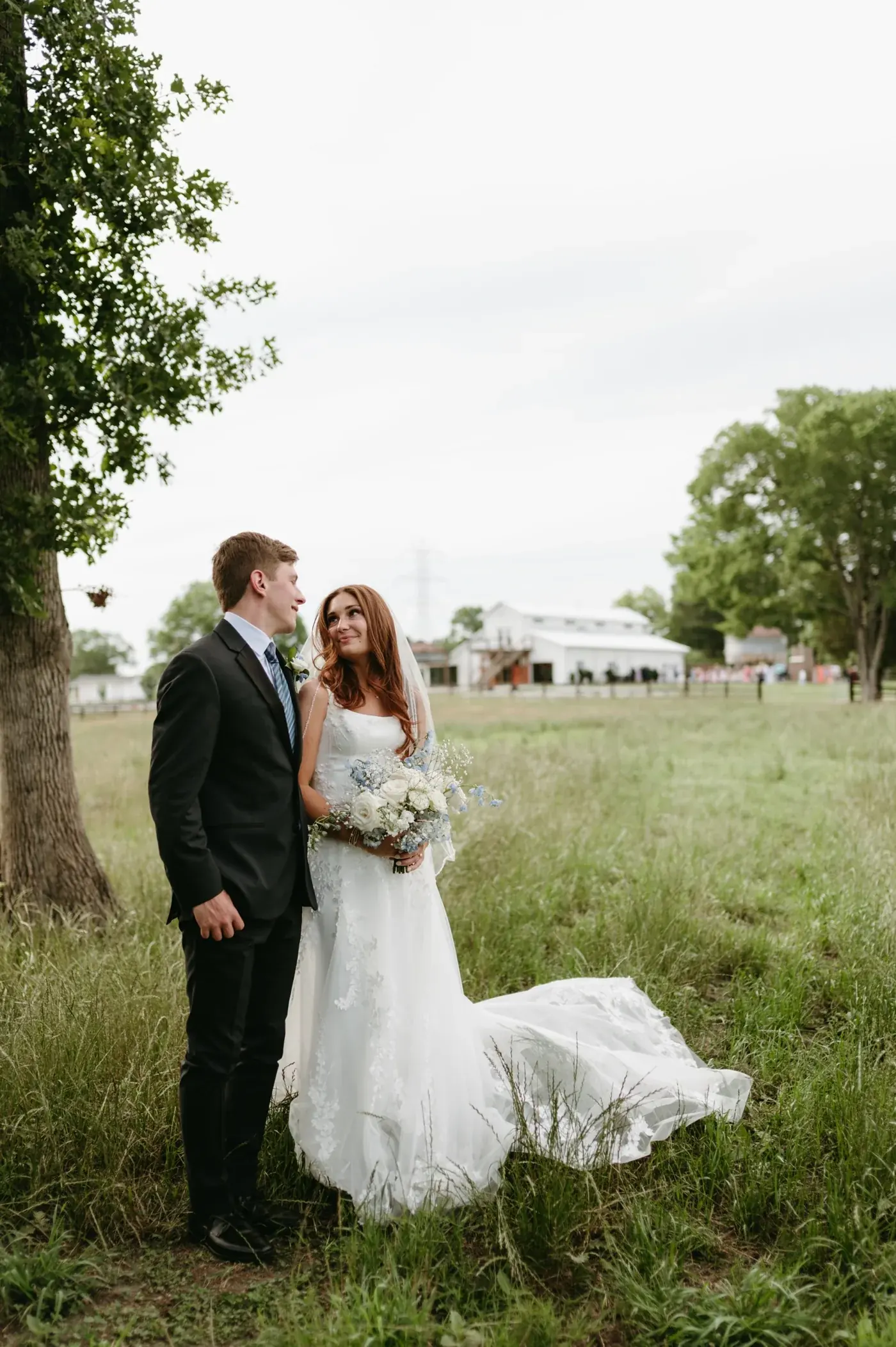 A bride and groom standing in tall grass beneath an oak tree, with the white barn and wedding guests across the pasture