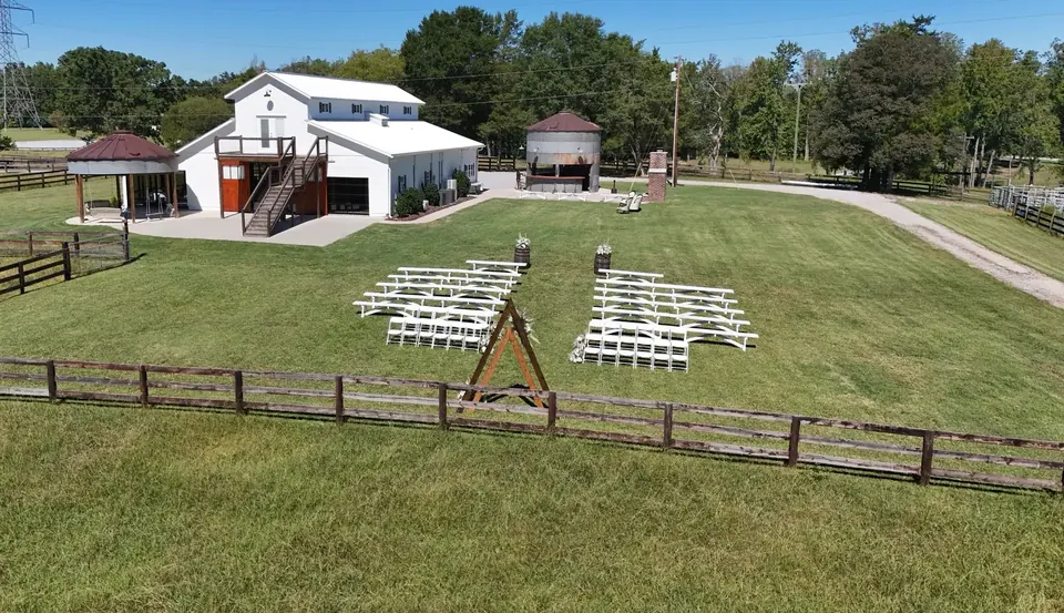 Aerial view across the fenced ceremony lawn toward the barn and Silo Bar, with benches and a wooden arch set for a wedding