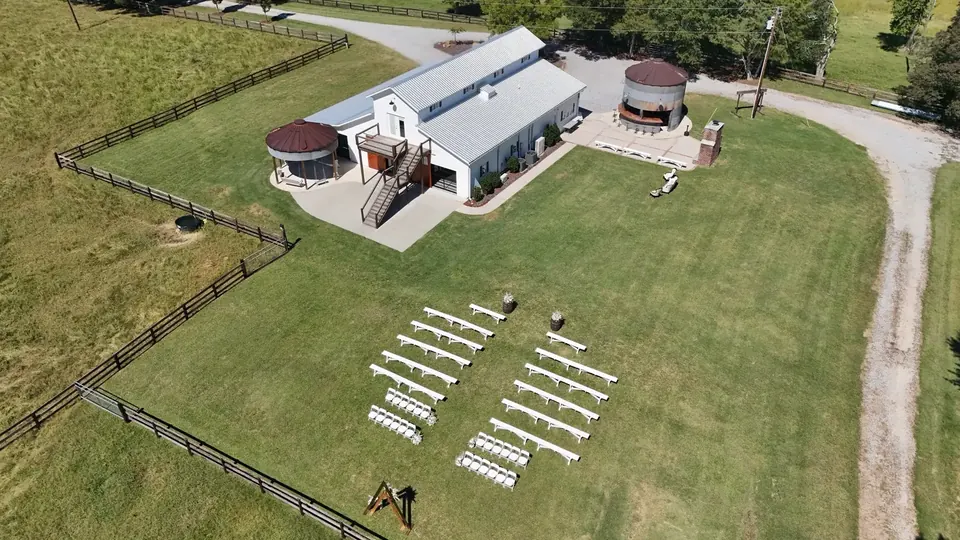 Aerial view of the white barn, two silos, patio, and ceremony lawn set with white benches and a wooden arch
