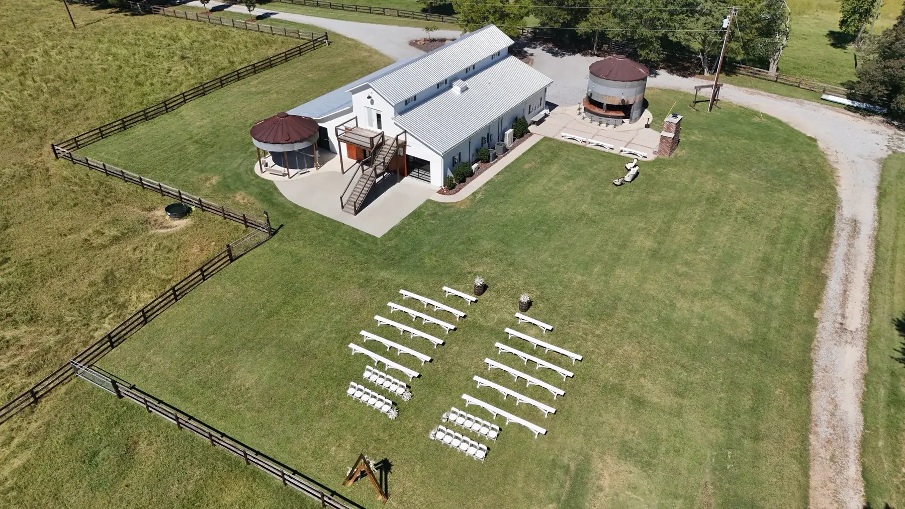 Aerial view of the white barn, two silos, patio, and ceremony lawn set with white benches and a wooden arch