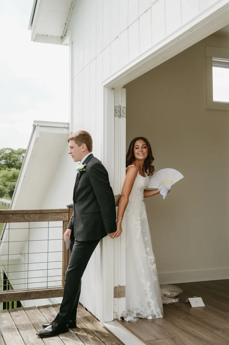 A groom on the white balcony and a smiling bride in the doorway holding hands around the door frame before the ceremony