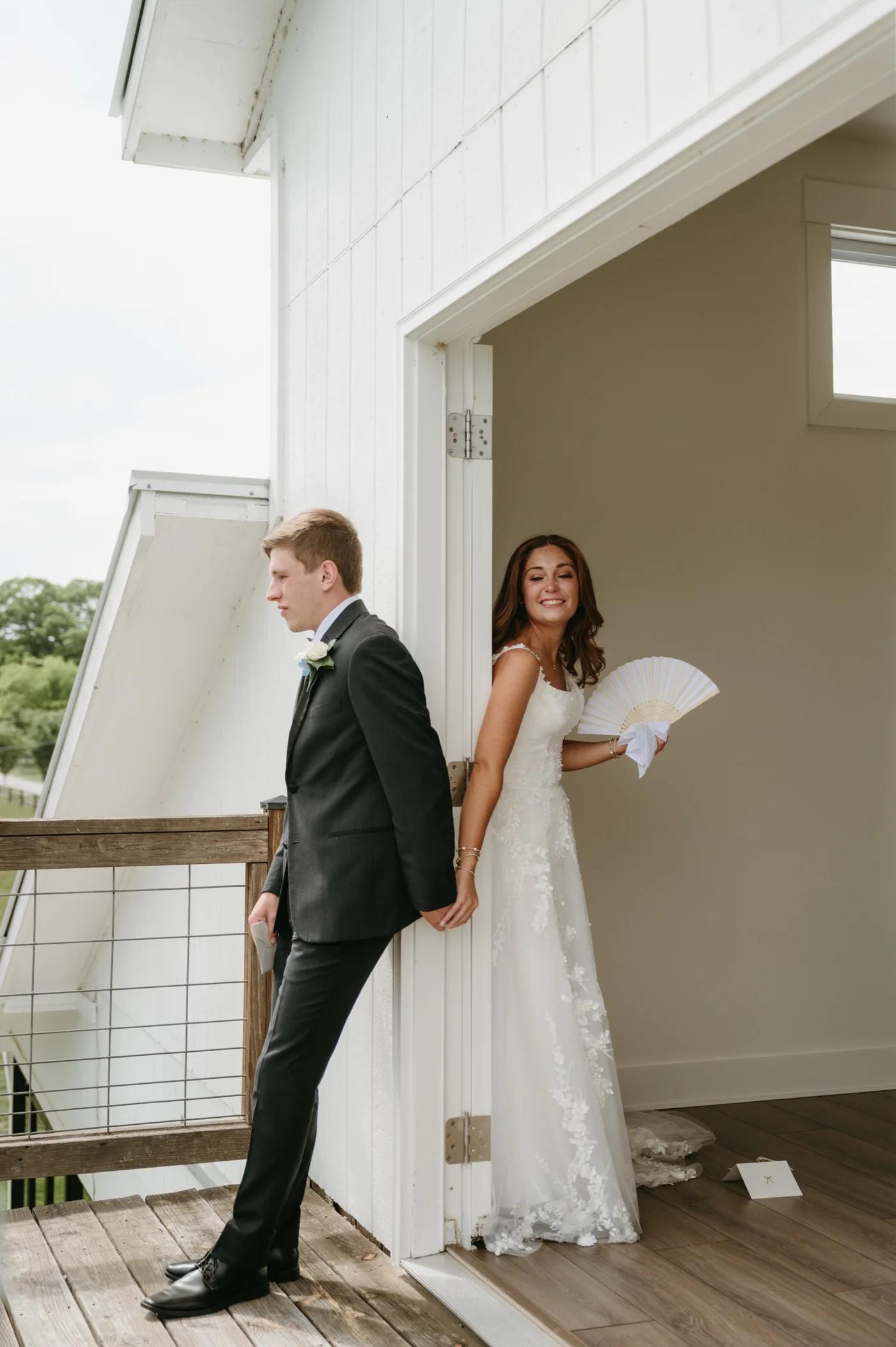 A groom on the white balcony and a smiling bride in the doorway holding hands around the door frame before the ceremony