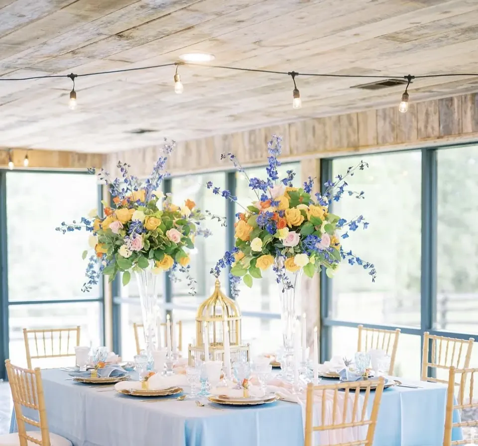 A blue-draped reception table with tall floral arrangements and gold-colored chairs beside the barn windows