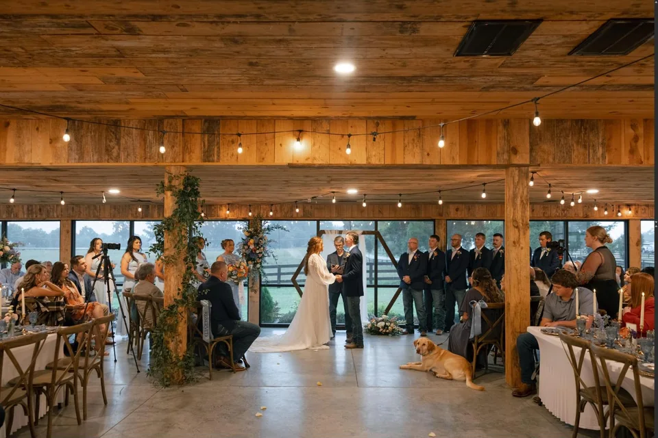 A wedding ceremony taking place inside the barn on a rainy day, guests seated at tables and the misty pasture visible through the glass wall
