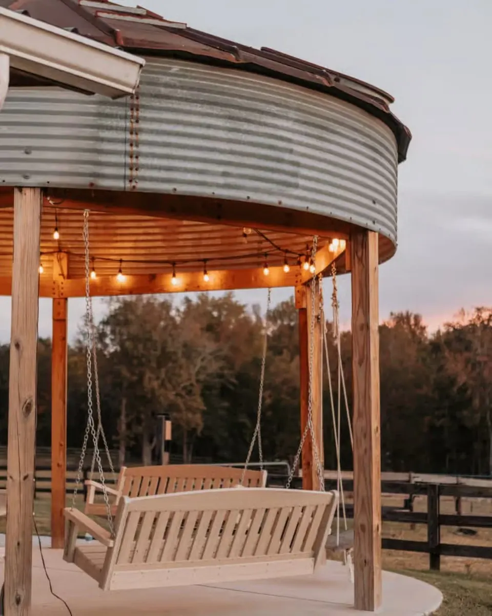 Wooden porch swings hanging inside the open silo beneath string lights at sunset, with the pasture fence beyond