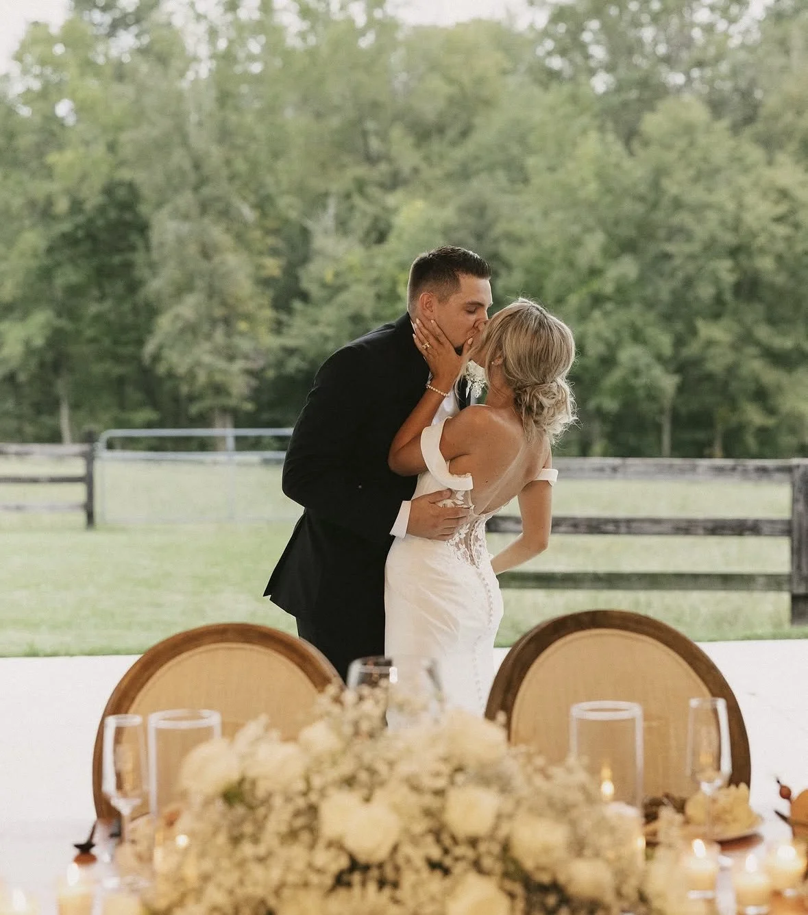 A bride and groom kissing behind a flower-covered sweetheart table, with the pasture fence and trees beyond