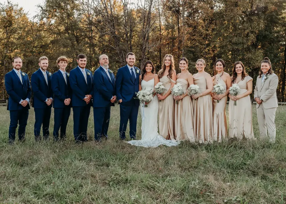 A wedding party in navy suits and champagne dresses standing in the tall grass of the pasture with autumn trees behind