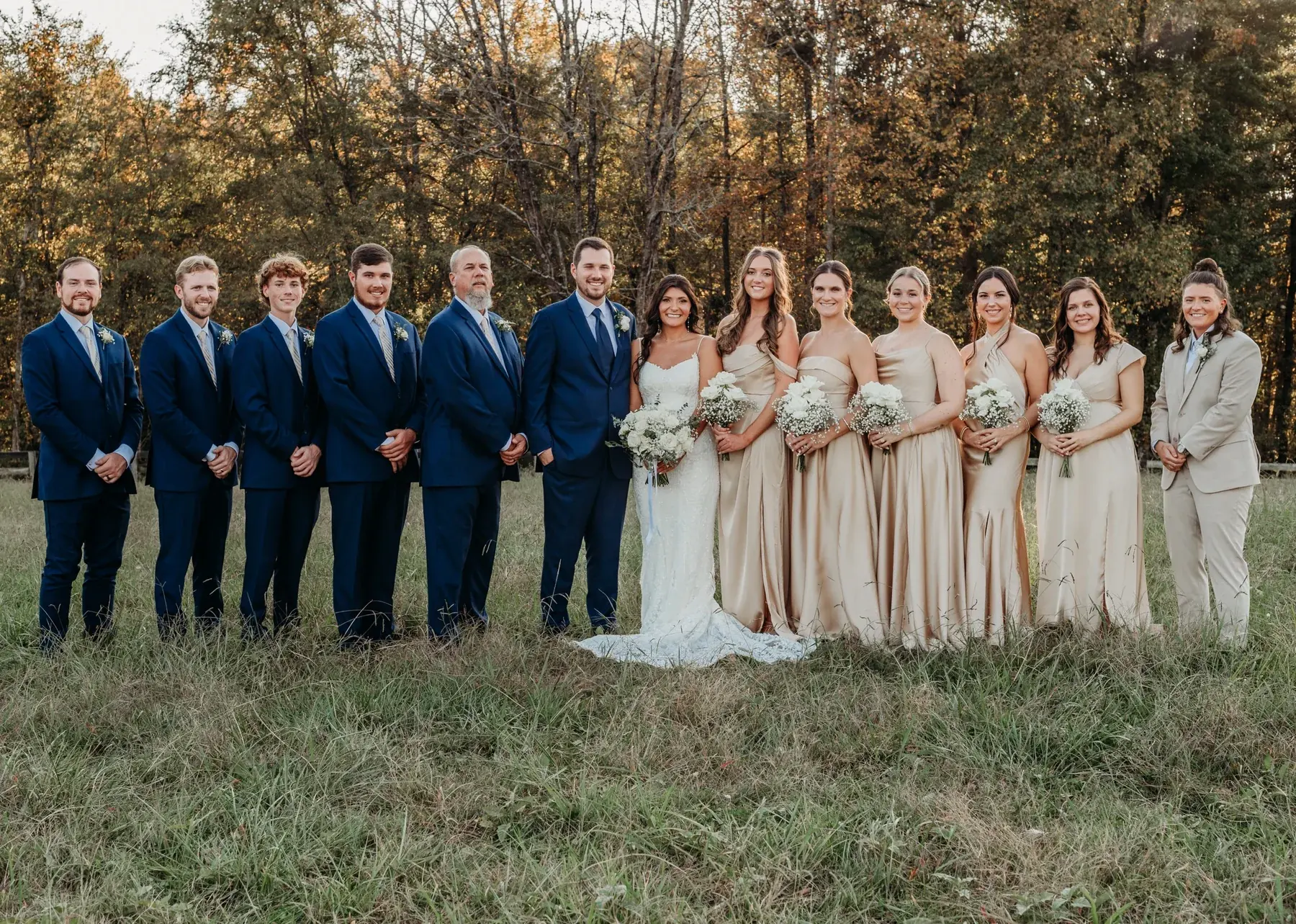 A wedding party in navy suits and champagne dresses standing in the tall grass of the pasture with autumn trees behind